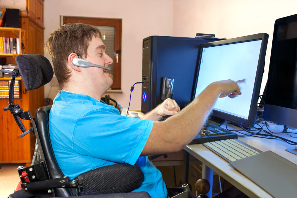 A young man with infantile cerebral palsy caused by a complicated birth sitting in a multifunctional wheelchair, using a computer with a wireless headset, reaching out to touch the touch screen.