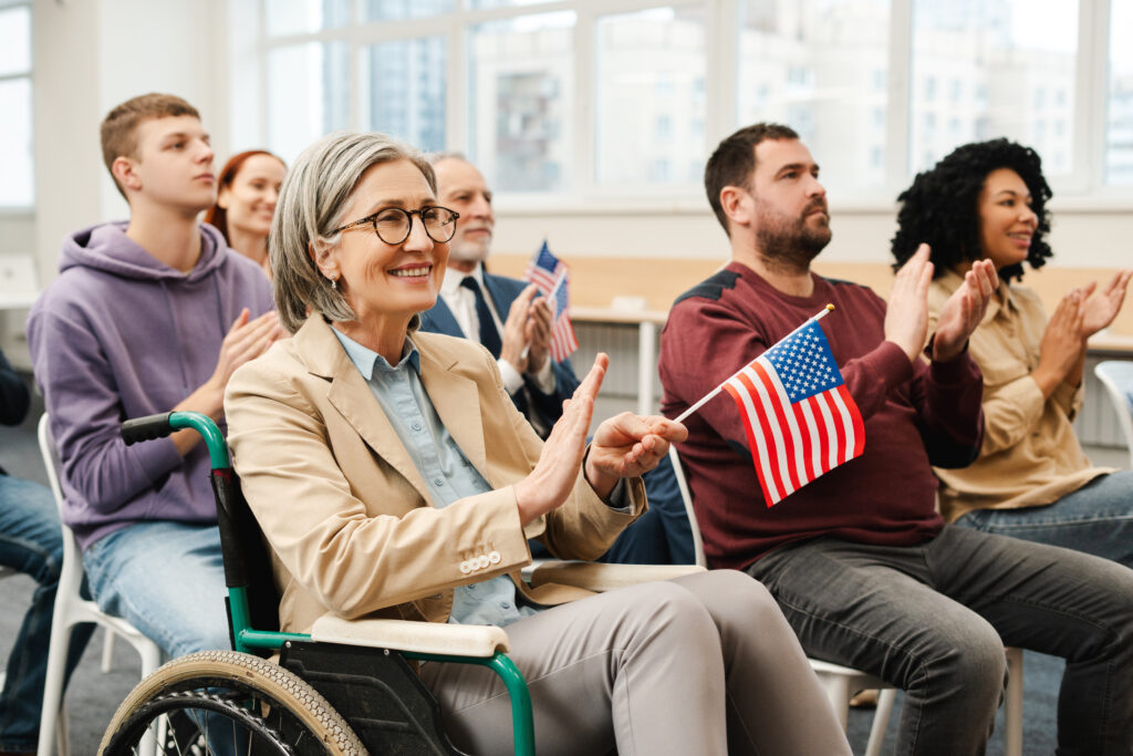 Group of multiracial naturalized US citizens at citizenship ceremony. A smiling senior woman in wheelchair holds an American flag.