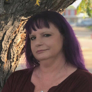 Image Description: Amber smiles at the camera. She is pictured from the shoulders up. Her hair is purple and falls straight behind her shoulder. She is wearing a burgundy top. Behind her is a tree trunk. 