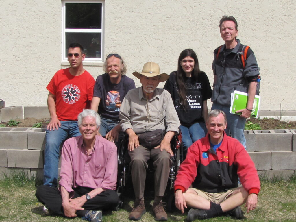 Ed Milewski with the CPWD Boulder Peer Group. Ed is seated in the middle in a wheelchair. he is wearing a tan shirt and brown pants, with a light tan hat. On the far left is a younger man wearing sunglasses and a red t-shirt. Next to him is an older gentleman with shoulder length blond hair and a mustache with sunglasses on his head, wearing a gray t-shirt with a graphic on it. Below in front is a gentlement with white hair, seated on the ground, wearing a rose-colored button up shirt. To the right of Ed, seated on the ground, is a man wearing a red and black jacket with grey hair. Standing behind the man, directly next to Ed is a young woman wearing a black t-shirt with long dark hair. To her right is another man standing, wearing a black and gray fleece, holding a green notebook. He has brown hair.