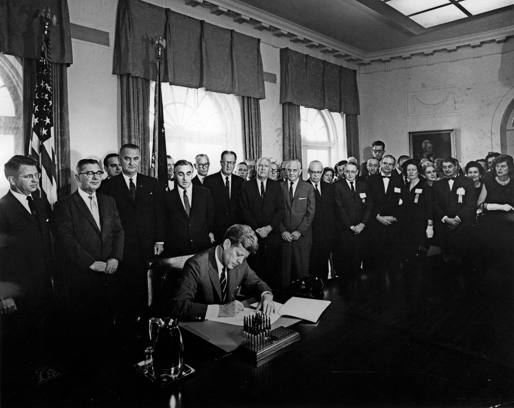 President John F. Kennedy signs the President's Panel into existence. Image description: The photo is in black and white. President Kennedy sits at a desk and is signing a document. He is wearing a dark suit and tie. Standing behind him are dozens of men and women in suits and dresses watching. Behind the crowd of men and women are large windows with paneled drapes.