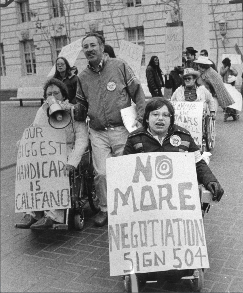 A black and white photo of demonstrators outside the San Fransisco HEW office building. In front of the group is Judy Heumann, a white woman who uses a wheelchair with short brown hair wearing glasses and a winter jacket with a “Sign 504” button pin. She is holding a sign that reads “NO MORE NEGOTIATION Sign 504.” To the left is Kitty Cone, a white woman who uses a wheelchair with short curly brown hair wearing glasses and a sweater. She is holding a sign that reads “OUR BIGGEST HANDICAP IS CALIFANO.” There are various other people around them holding signs and a mega phone.