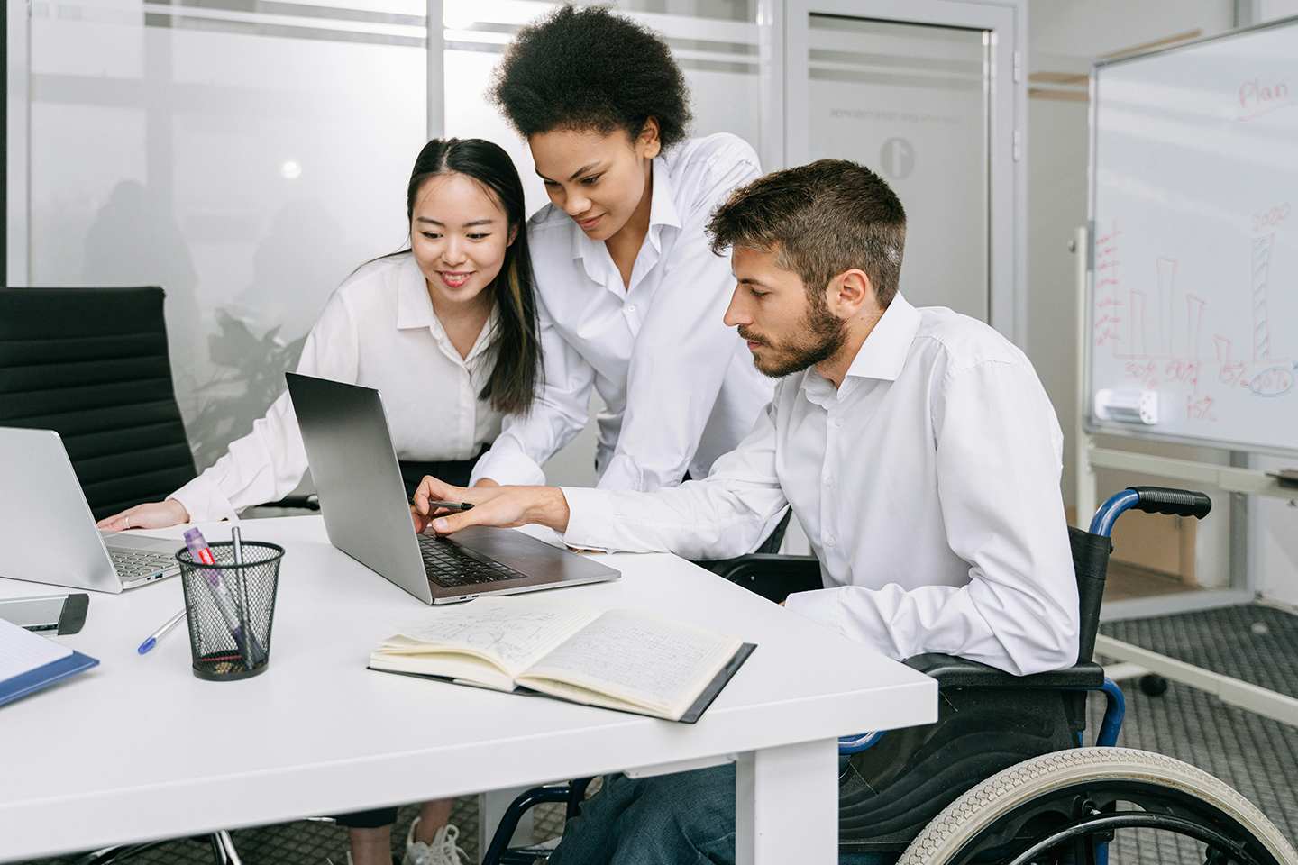 Three people stand over a desk looking at a laptop. The man on the right is a wheelchair user.