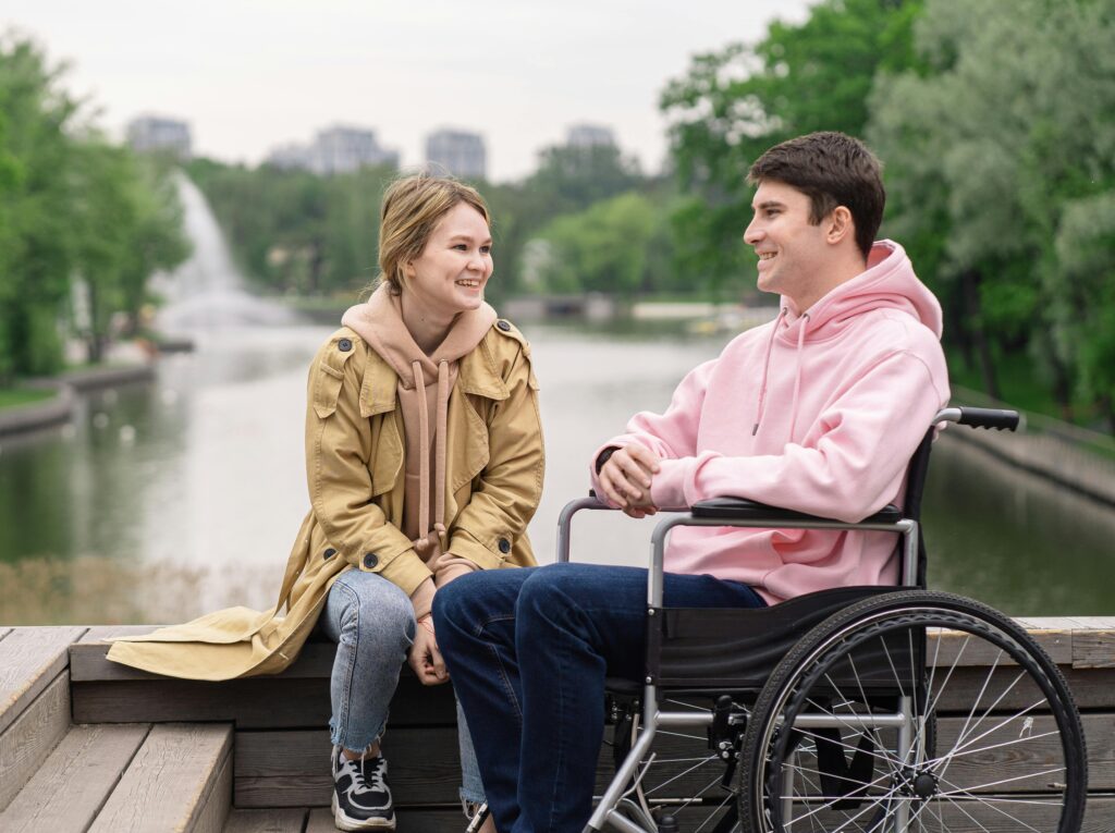 A man who is a wheelchair user sits outside with a woman by a lake. They are smiling at one another.